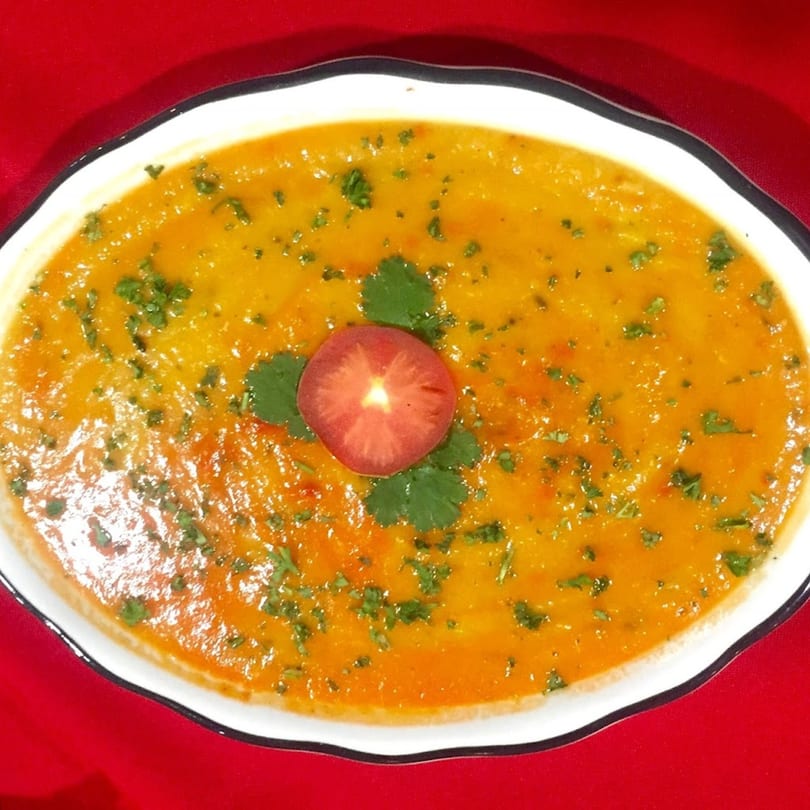 A vibrant orange Indian curry, garnished with fresh green herbs and a tomato slice, in a white dish.
