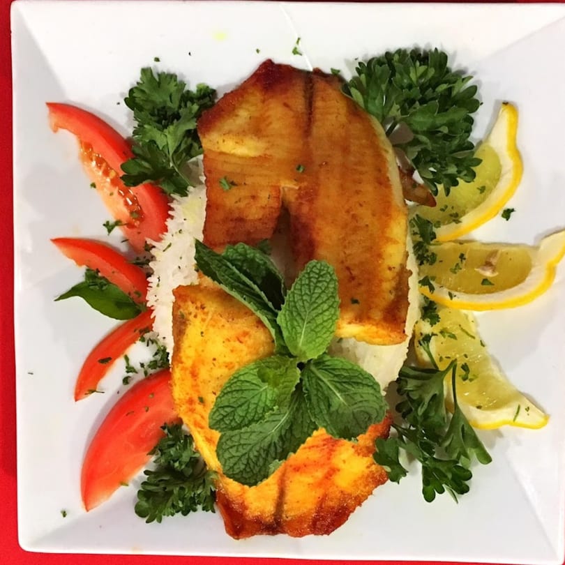 Golden-brown fish fillets on a bed of rice, garnished with tomatoes, lemon slices, parsley, and mint.