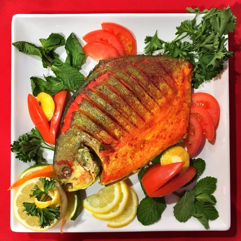 Whole fried fish, garnished with lemon, lime, tomatoes, and fresh herbs on a white plate.