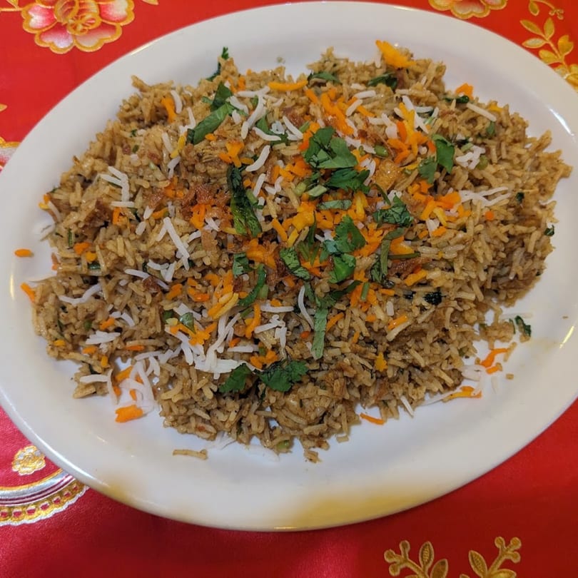 A white plate of Indian biryani rice, garnished with cilantro and shredded carrots, on a red tablecloth.